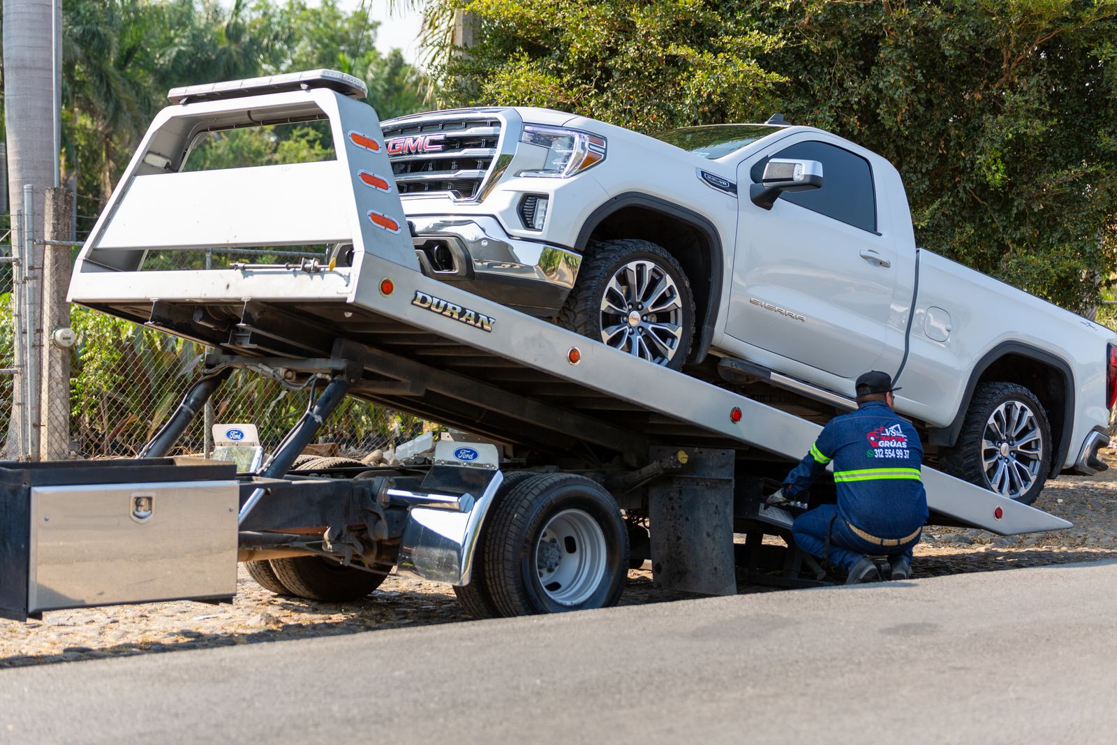 A white GMC pickup truck being winched onto the tilted bed of a flatbed tow truck while a worker secures it.