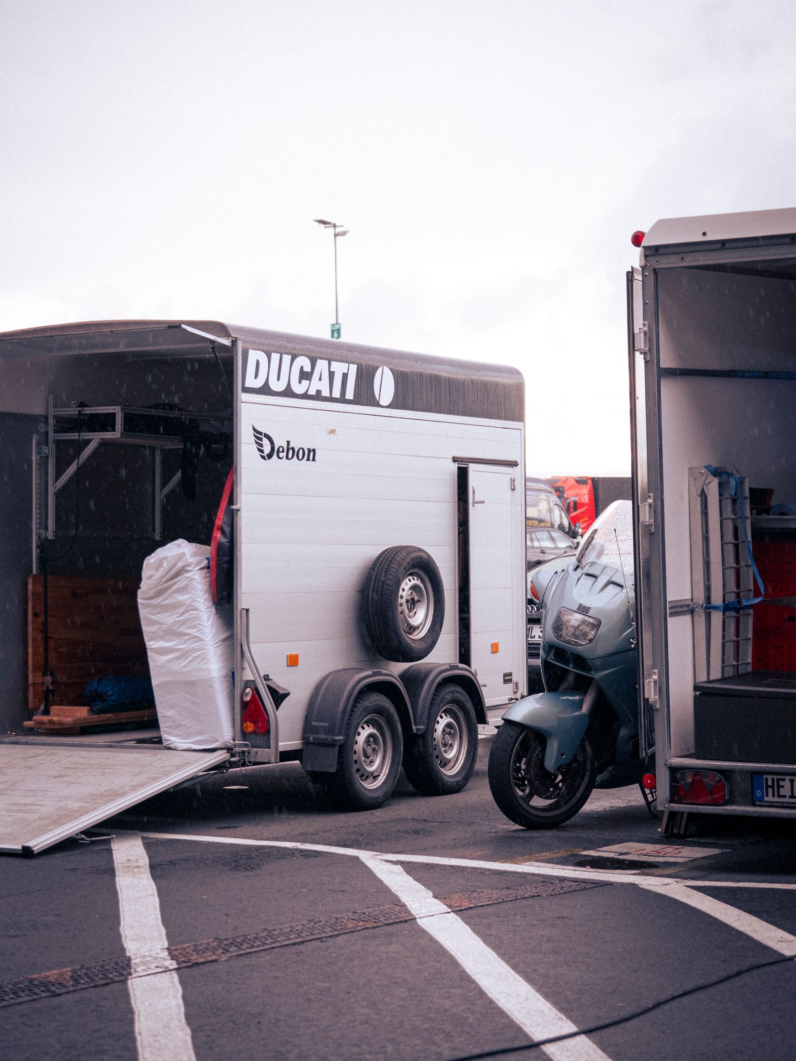 A motorcycle standing beside the open rear doors of an enclosed Ducati transport trailer, with a spare wheel and ramp inside.